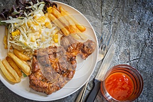 Grilled pork steaks on white plate with salad and french fries and ketchup on stone background