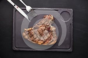 Grilled ribeye beef steak with fork and knife on stone plate on black background