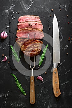 Grilled ribeye beef steak medium rare on the fork, black stone background. Top view, flat lay, copy space