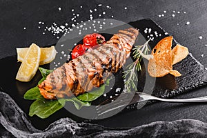 Grilled salmon fillet garnished with spinach, lemon, herbs on plate over wooden background. Hot fish dish