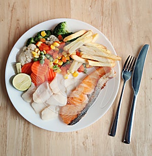 Grilled salmon fish and baked cod fish decorated with various vegetables corn carrot peas lemon french fries and sauces