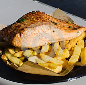 Grilled salmon fish with chips on a black plastic plate.