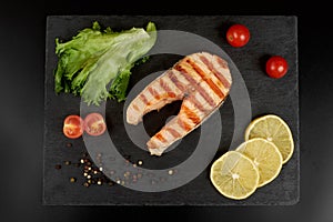 Grilled salmon steak with a lemon, tomatoes and salad on a slate plate.