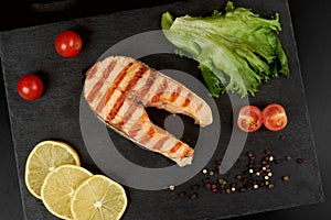 Grilled salmon steak with a lemon, tomatoes and salad on a slate plate.