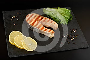 Grilled salmon steak with a lemon, tomatoes and salad on a slate plate.