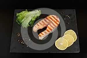 Grilled salmon steak with a lemon, tomatoes and salad on a slate plate.