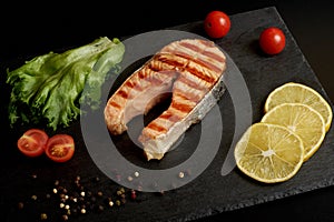 Grilled salmon steak with a lemon, tomatoes and salad on a slate plate.