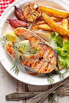 Grilled salmon steak, a portion of grilled salmon with fresh lettuce and potato wedges on a white ceramic plate on a wooden rustic