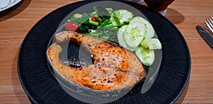 Grilled salmon steak with slices cucumber, tomato and fresh vegetable on black dish or plate on wooden table