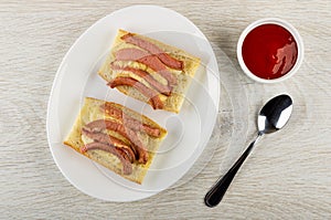 Grilled sandwiches from bread with sausages and cheese in plate, bowl with ketchup, spoon on table. Top view