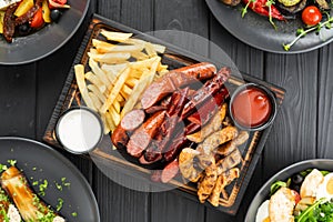 grilled sausages french fries ketchup and mayonnaise sauce on cutting board on wooden background, top view