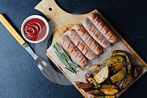 Grilled sausages on a wooden chopping Board. Fried potatoes, rosemary, tomato ketchup. Unhealthy diet. Dark background