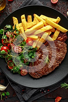 Grilled sirloin steak with potato fries and vegetables, tomato salad in a black plate. rustic table