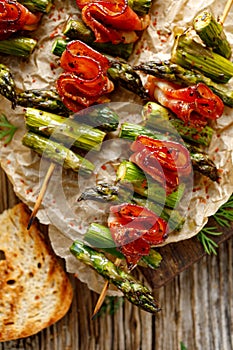 Grilled skewers of green asparagus and bacon on a wooden table, top view.