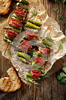 Grilled skewers of green asparagus and bacon on a wooden table, top view.