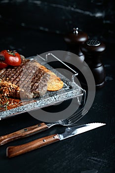 Grilled steak with knife and fork carved on black stone slate. Steak on a hot marble stone. Copy space, dark background