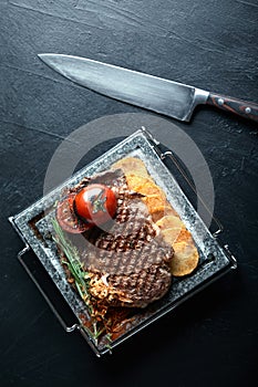 Grilled steak with knife and fork carved on black stone slate. Steak on a hot marble stone. Copy space, dark background