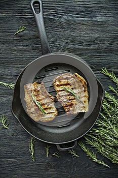 Grilled steak on a round grill pan, garnished with spices for meat, rosemary, greens and vegetables on a dark wooden background
