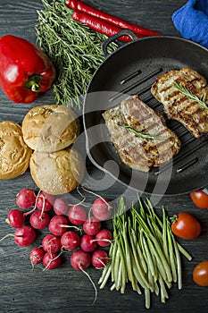 Grilled steak on a round grill pan, garnished with spices for meat, rosemary, greens and vegetables on a dark wooden background