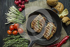 Grilled steak on a round grill pan, garnished with spices for meat, rosemary, greens and vegetables on a dark wooden background