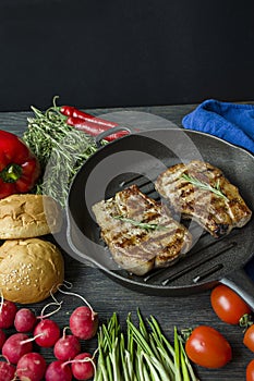 Grilled steak on a round grill pan, garnished with spices for meat, rosemary, greens and vegetables on a dark wooden background