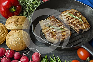 Grilled steak on a round grill pan, garnished with spices for meat, rosemary, greens and vegetables on a dark wooden background