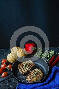 Grilled steak on a round grill pan, garnished with spices for meat, rosemary, greens and vegetables on a dark wooden background.