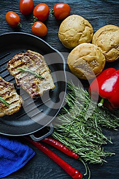 Grilled steak on a round grill pan, garnished with spices for meat, rosemary, greens and vegetables on a dark wooden background