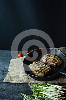 Grilled steak on a round grill pan, garnished with spices for meat, rosemary, greens and vegetables on a dark wooden background.