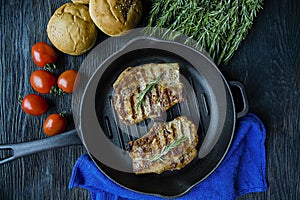 Grilled steak on a round grill pan, garnished with spices for meat, rosemary, greens and vegetables on a dark wooden background