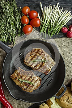 Grilled steak on a round grill pan, garnished with spices for meat, rosemary, greens and vegetables on a dark wooden background