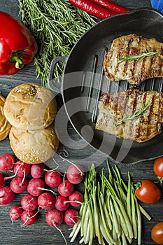 Grilled steak on a round grill pan, garnished with spices for meat, rosemary, greens and vegetables on a dark wooden background