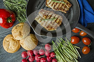 Grilled steak on a round grill pan, garnished with spices for meat, rosemary, greens and vegetables on a dark wooden background