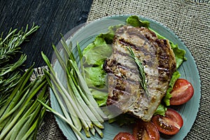 Grilled steak served on a plate, decorated with spices for meat, rosemary, greens and vegetables on a dark wooden background