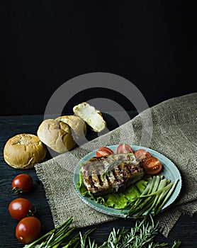 Grilled steak served on a plate, decorated with spices for meat, rosemary, greens and vegetables on a dark wooden background.