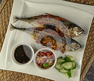 Grilled Torpedo Scad fish Ikan Cencaru Bakar in Malay with hot soy sauce, hot tamarind juice and fresh cucumber on white plate