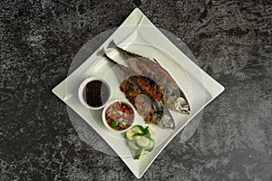 Grilled Torpedo Scad fish Ikan Cencaru Bakar in Malay with hot soy sauce, hot tamarind juice and fresh cucumber on white plate