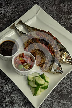 Grilled Torpedo Scad fish Ikan Cencaru Bakar in Malay with hot soy sauce, hot tamarind juice and fresh cucumber on white plate
