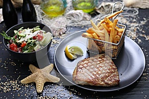 Grilled tuna steak served with potato fries in a metal serving basket and salad mix, on black plates, selective focus.