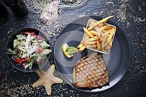 Grilled tuna steak served with potato fries in a metal serving basket and salad mix, on black plates, top view.