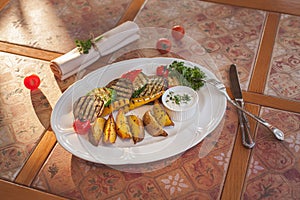 Grilled vegetables with baked potatoes on an oval white plate, lit by sunlight. with sauce and herbs lying next to a fork and a