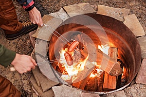 Grilling sausages over a campfire, campers roasting sausages on toasting forks. Fire place, friends, tourists are