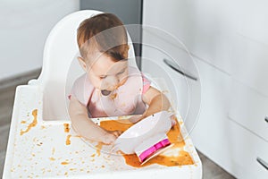grimy dirty baby 9 months old sits in a highchair and eats complementary foods vegetable puree soup spilled from a plate, the