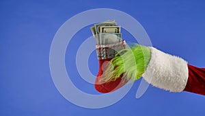 The Grinch's green haired hand hold a Christmas sock with dollar bills on a blue isolated background. Gift Snatcher