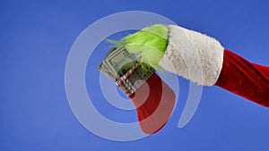 The Grinch's green haired hand hold a Christmas sock with dollar bills on a blue isolated background. Gift Snatcher