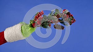 The Grinch's green haired hand hold a decorative fir tree branch with pinecones and red berries. Gift Snatcher