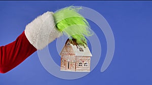 The Grinch's green haired hand hold out a decorative snowy house on a blue isolated background. Gift Snatcher