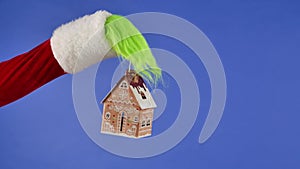 The Grinch's green haired hand holds a decorative snowy house on a blue isolated background. Gift Snatcher Cosplay