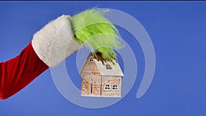 The Grinch's green haired hand holds a decorative snowy house on a blue isolated background. Gift Snatcher Cosplay