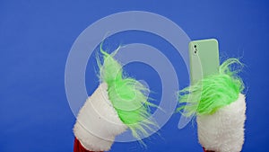 The Grinch's green haired hand holds smartphone and shows a thumbs up gesture. Gift kidnapper cosplay. Christmas and photo
